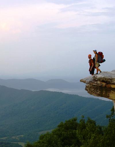 A person stands on McAfee Knob — a piece of land jutting out over a valley with the rolling Blue Ridge Mountains in the background — off the Appalachian Trail